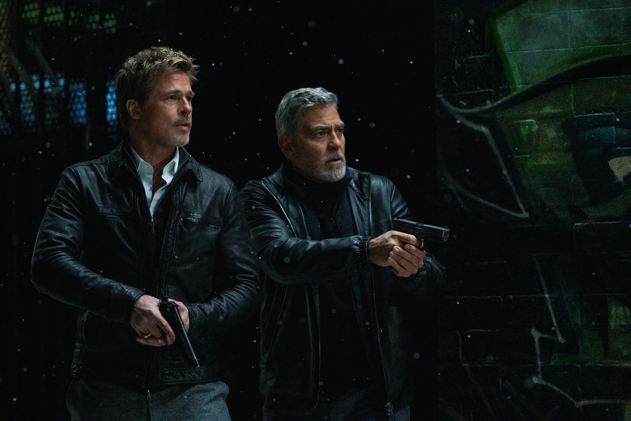 George Clooney and Brad Pitt stand side by side holding guns.