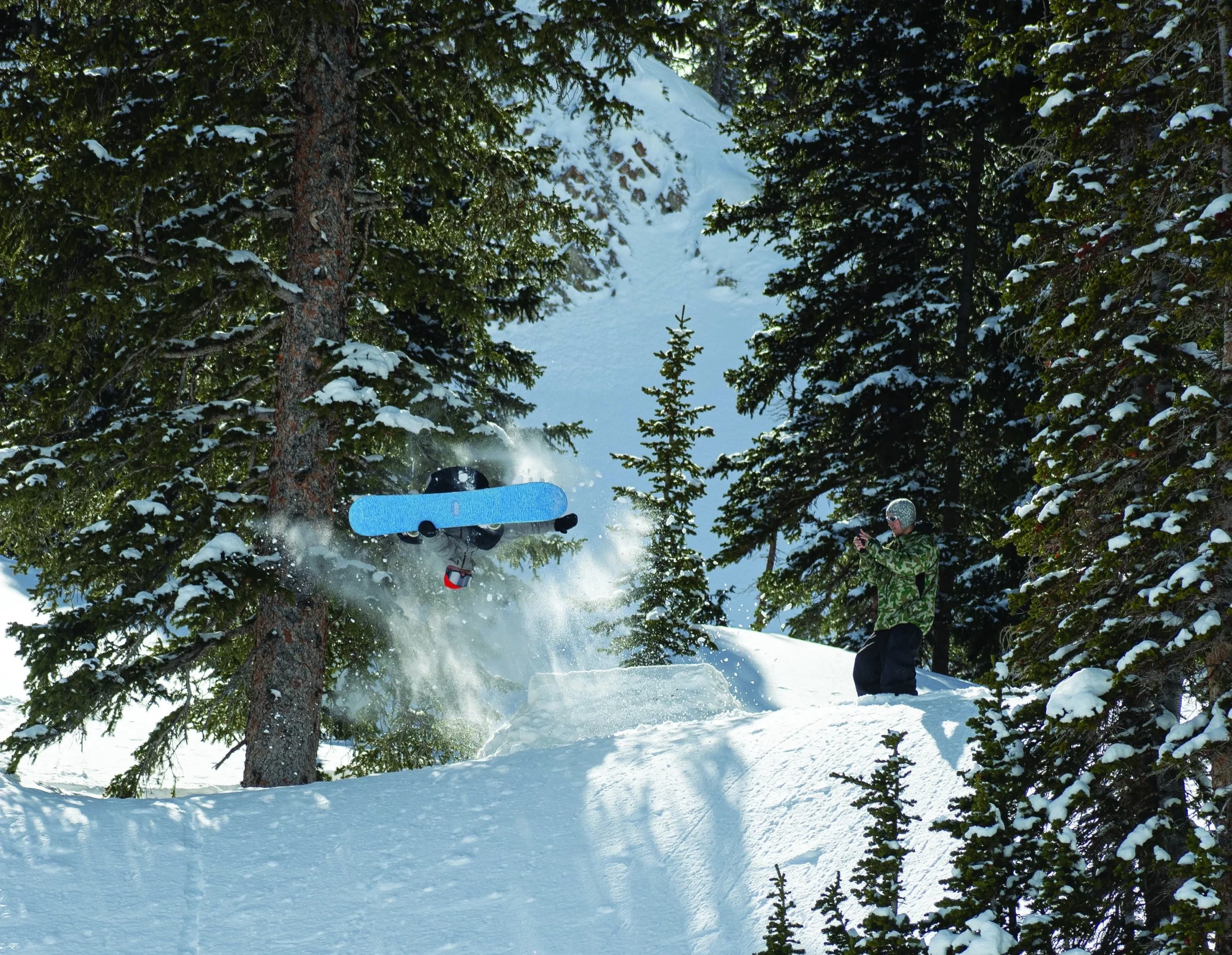 Two snowboarders in the mountains, one is filming while the other is taking a jump off of the hill.