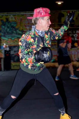 Eliza James wears black boxing gloves to throw some practice punches in front of a wall covered in bright graffiti.