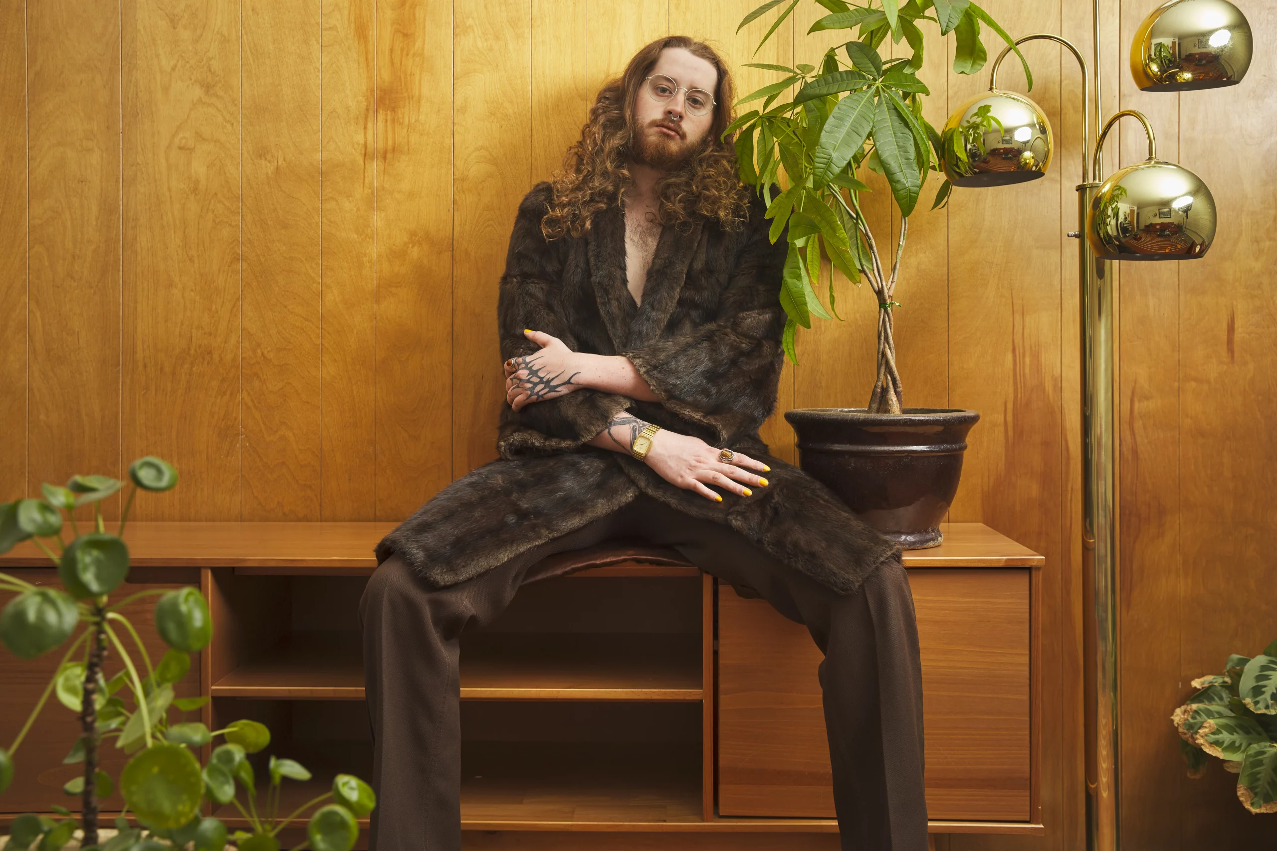Dom Ducote sits against a wooden wall with plants and a lamp behind him.