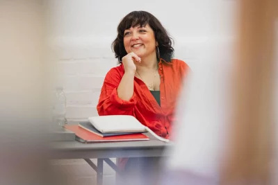 A woman in an orange blouse sits at a table with a plastic water bottle and notebooks on it. 