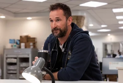 Noah Wyle stands at desk.