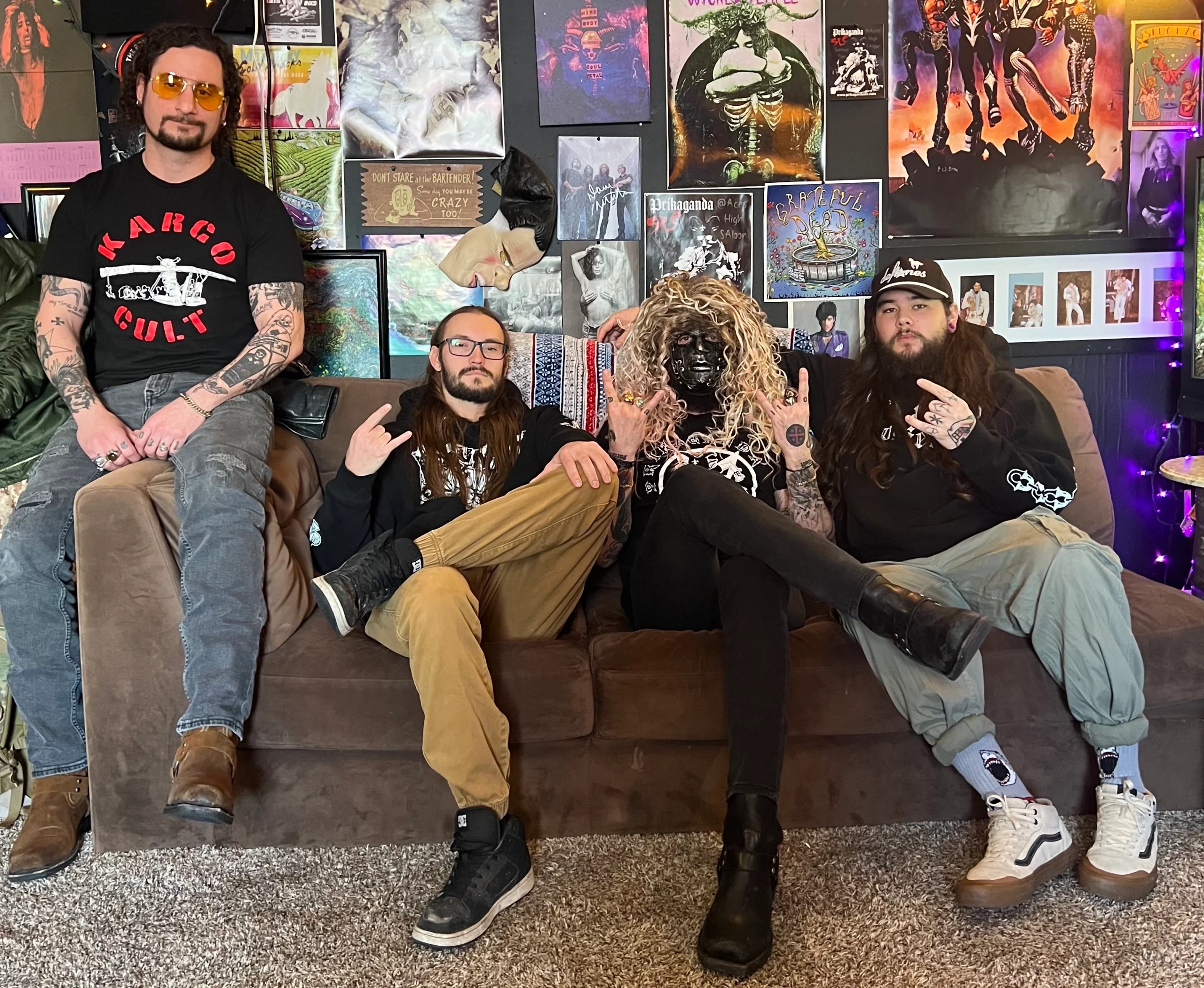The members of Wicked Temple, one of which wears a wig and mask, sit on a couch in front of a wall plastered with posters.