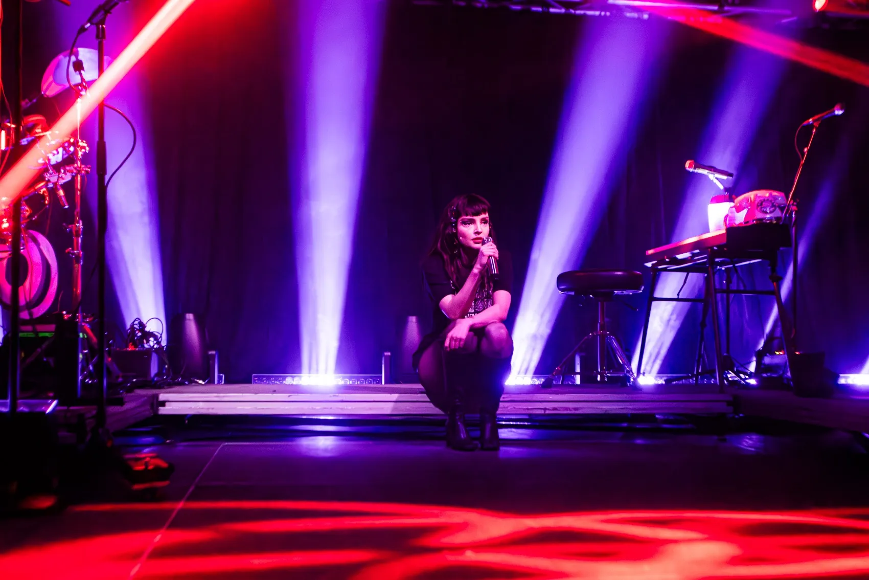 Lauren Mayberry sits at the edge of the stage, illuminated by red and purple lights.