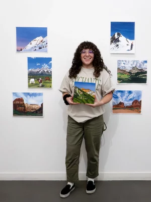 Steph Alpizar stands and smiles with her paintings on the wall surrounding her. 