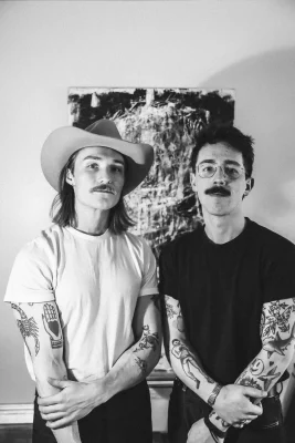 Two men stand in front of a poster in a black and white photo.