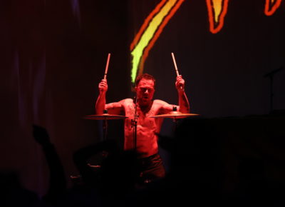 SOFT PLAY's Isaac Holman, hands up with drum sticks in hand, bathed in red stage lights with the main room in darkness. Photo: Logan Sorenson.