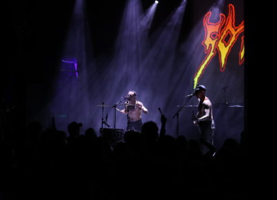 Isaac Holman of SOFT PLAY, on stage with band mate Laurie Vincent with some back lighting kissing the drumsticks in hands and packed audience just feet away. Photo: Logan Sorenson.