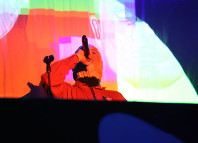 DJ Próvaí holding the mic, at the back of stage, beaming in the colors of the stage lights. Photo: Logan Sorenson.