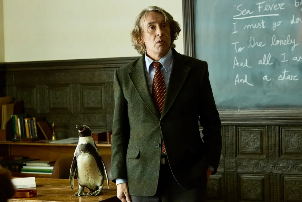 Steve Coogan stands next to a penguin standing on a desk.