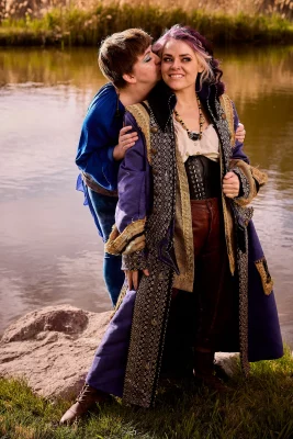 One theatre actor kisses another on the cheek by a river.