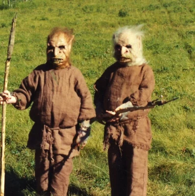 Two men in goblin masks and burlap shirts stand in a field