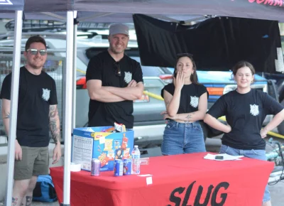 Team members at a SLUG tabling booth under a tent.